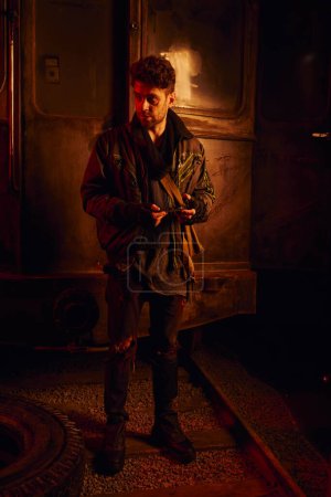 Photo for Man in dirty clothes with spare part of rusty carriage in post-disaster subway with red light - Royalty Free Image