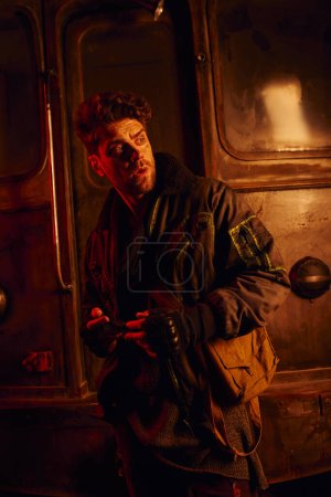 homme inquiet se sentant en danger et regardant loin près de chariot rouillé dans le métro post-catastrophe dans la lumière rouge