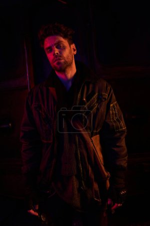 Photo for Grim man in black worn jacket looking at camera in dark post-apocalyptic subway with red light - Royalty Free Image