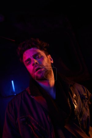 exhausted unshaven man looking at camera in dark post- apocalyptic subway with neon light