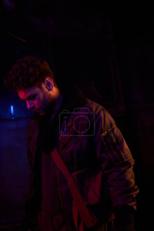 Photo for Hopeless unshaven man in worn clothes standing on darkness of post-disaster subway with neon light - Royalty Free Image