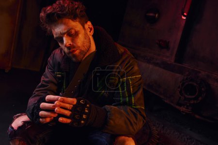 unshaven man in fingerless gloves holding military knife in post-disaster underground with red light