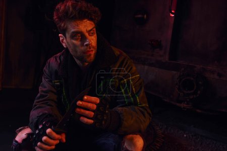 Foto de Hombre reflexivo en guantes sin dedos sentado con cuchillo militar en la oscuridad subterránea con luz roja - Imagen libre de derechos
