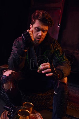 hungry man in worn outfit opening canned food with military knife in dark post-disaster underground Mouse Pad 675366170