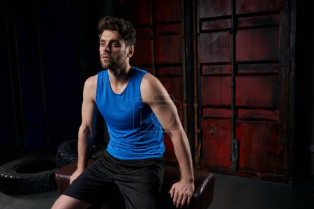 Photo for Unshaven man in sportswear sitting on sport bench at night on urban street, outdoor workout - Royalty Free Image