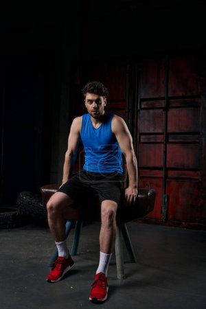 Photo for Confident man in sportswear looking at camera and sitting on sport bench at night, full length - Royalty Free Image