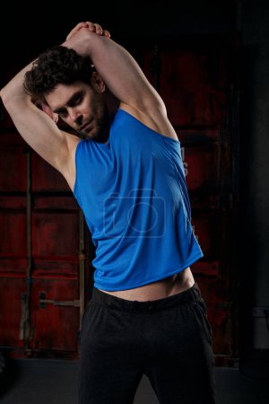athletic man in blue tank top training and stretching arms in dark city at night, street fitness
