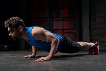 Photo for Concentrated man in blue tank top training in plank pose in darkness of night urban street - Royalty Free Image