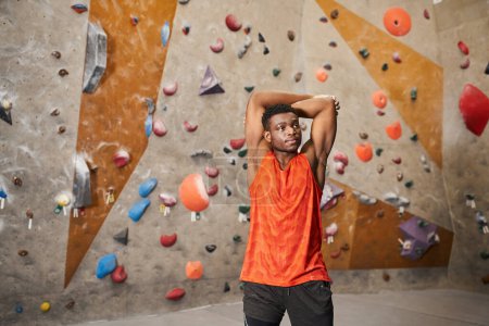 sporty african american man with hands behind head looking away with climbing wall on backdrop