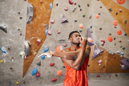 athletischer afrikanisch-amerikanischer Mann im orangefarbenen Hemd beim Aufwärmen vor dem Klettern an der Wand, Klettern