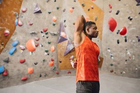 afrikanisch-amerikanischer athletischer Mann im orangefarbenen Hemd, der sich mit den Händen hinter dem Rücken aufwärmt, Kletterkonzept