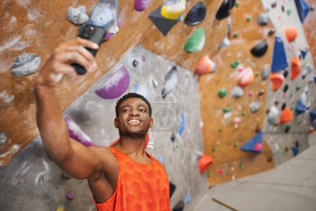 happy african american man taking selfie and smiling cheerfully with climbing wall backdrop