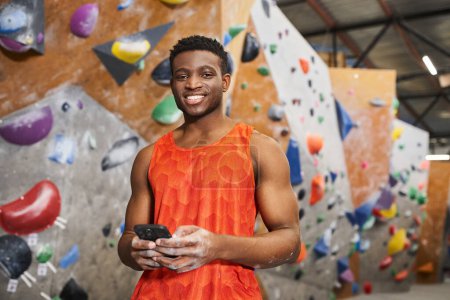 jolly africano americano hombre en naranja camisa celebración de teléfono móvil y mirando a la cámara, escalada en roca