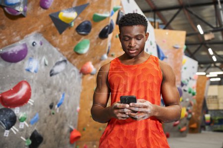 Konzentrierter afrikanisch-amerikanischer Mann in orangefarbenem Hemd blickt auf sein Handy, klettert