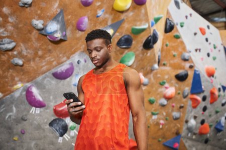 hombre afroamericano enfocado mirando su teléfono móvil con atención con el fondo de la pared de escalada