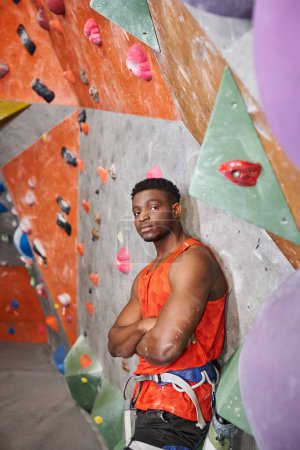 Afrikanisch-amerikanischer athletischer Mann in orangefarbenem Hemd nahe der Kletterwand mit verschränkten Armen auf der Brust