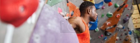 hombre afroamericano guapo con camisa naranja al lado de la pared de escalada mirando hacia otro lado, pancarta