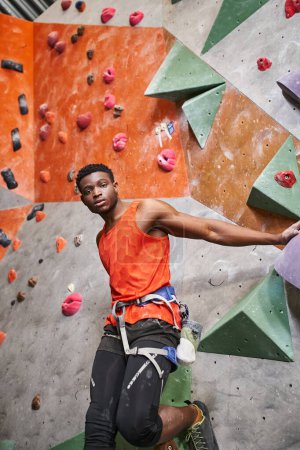 handsome african american man with alpine harness in orange shirt posing next to rock climbing wall