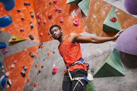 athletic african american male model with alpine harness posing next to rock climbing wall