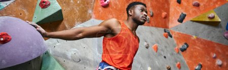 handsome african american man with alpine harness posing near climbing wall and looking away, banner