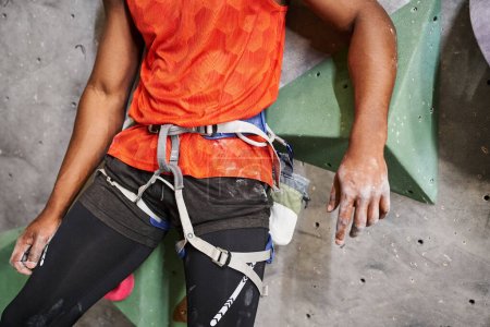 abgeschnittene Ansicht eines muskulösen afrikanisch-amerikanischen Mannes mit orangefarbenem Hemd und Alpingeschirr, Bouldern