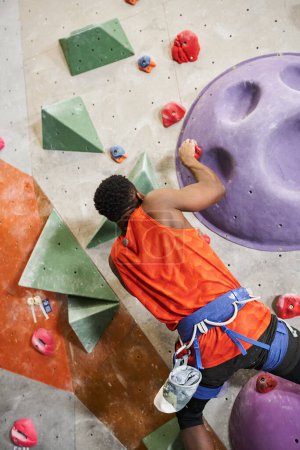 vertical shot of athletic african american man with alpine harness climbing up rock wall, bouldering