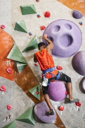 vertical shot of muscular african american man with alpine harness climbing up rock wall, bouldering