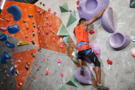 sporty african american man in orange shirt with alpine harness climbing up wall, sport concept