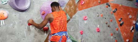 sporty african american man in orange shirt climbing up rock wall with alpine harness, banner