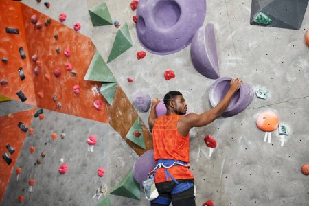 Photo for Good looking african american man posing and climbing up rock wall with alpine harness, sport - Royalty Free Image