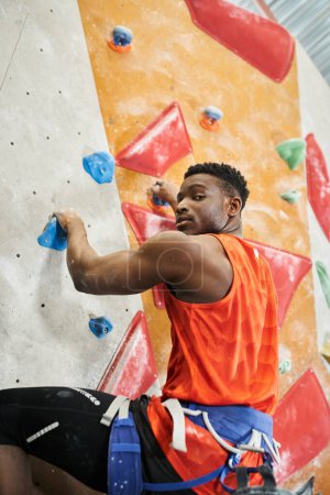 Photo for Vertical shot of handsome african american man with alpine harness climbing and looking at camera - Royalty Free Image