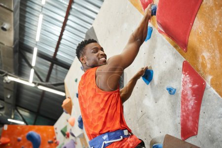 cheerful african american man smiling happily at camera while climbing up rock wall, bouldering