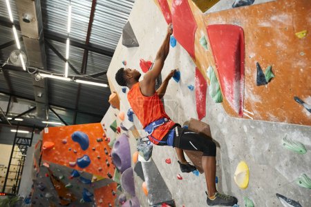 guapo afroamericano hombre de camisa naranja posando de perfil mientras trepa por la pared de roca