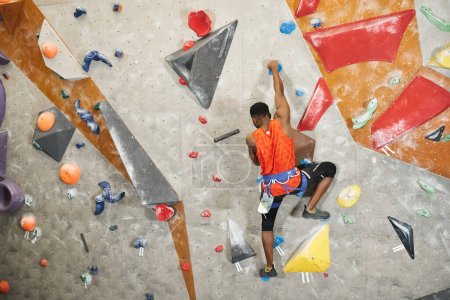 back view of sporty african american man with alpine harness climbing up rock wall, bouldering