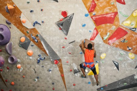 back view of muscular african american man with alpine harness in orange shirt climbing up wall