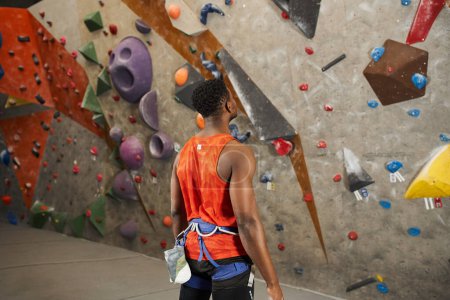sporty african american man in orange shirt with alpine harness ready to climb rock wall, back view