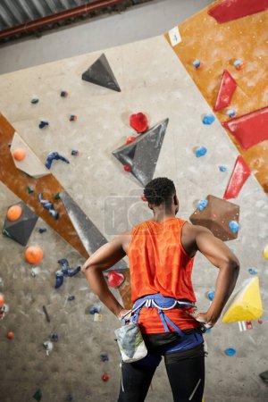 vista posterior del hombre americano africano deportivo en camisa naranja con arnés alpino con manos akimbo