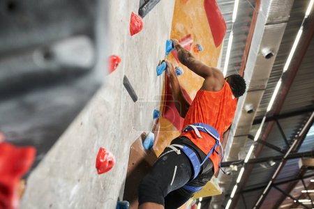 muscular african american man climbing up rock wall with alpine harness, bouldering concept