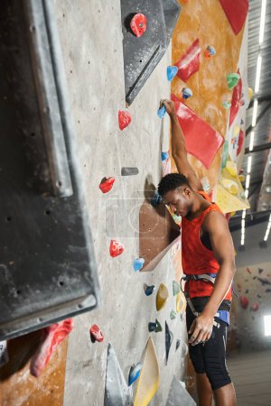 Foto de Plano vertical del hombre afroamericano colgando de sus dedos en la pared de roca y mirando hacia abajo - Imagen libre de derechos