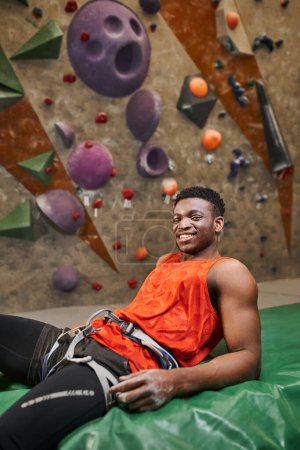 vertical shot of jolly african american relaxing on bouldering pad and smiling cheerfully at camera