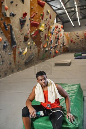 hombre americano africano deportivo que se relaja en la almohadilla de choque después de escalar pared de roca, toalla y botella de agua