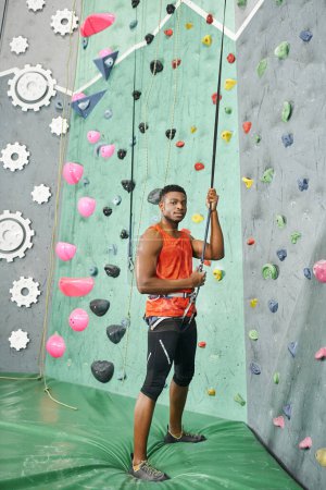 Photo for Vertical shot of african american man in orange shirt using climbing rope on rock wall, bouldering - Royalty Free Image
