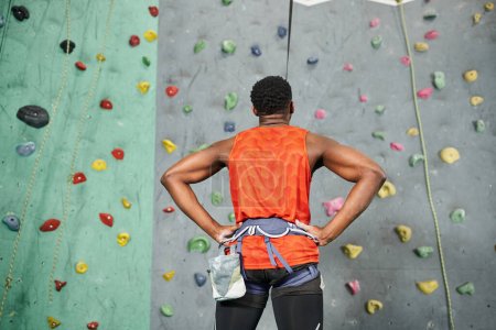 Rückenansicht des sportlichen afrikanisch-amerikanischen Mannes, der bereit ist, mit den Händen auf den Hüften die Boulderwand hinaufzuklettern
