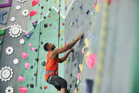 Photo for Young african american man climbing up bouldering wall with safety rope and alpine harness - Royalty Free Image