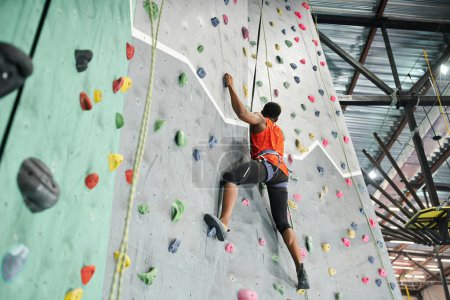 sporty african american man in orange shirt climbing up bouldering wall gripping strongly on rocks