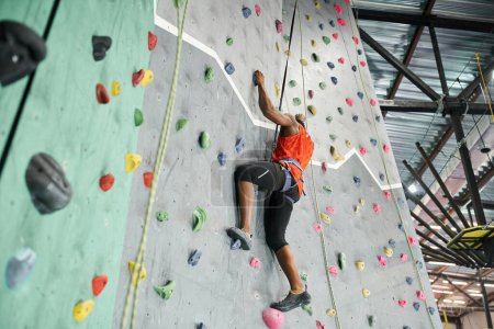 Photo for Strong athletic african american man with alpine harness climbing up rock wall gripping on rocks - Royalty Free Image