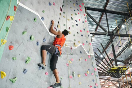 Photo for Strong african american man in orange shirt using safety rope and alpine harness to climb up wall - Royalty Free Image