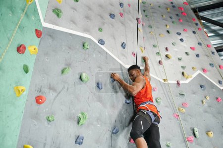 Photo for Sporty young african american man using safety rope and harness to climb up rock wall, bouldering - Royalty Free Image