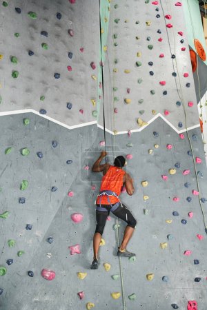 back view of athletic african american man with alpine harness and safety rope climbing up rock wall