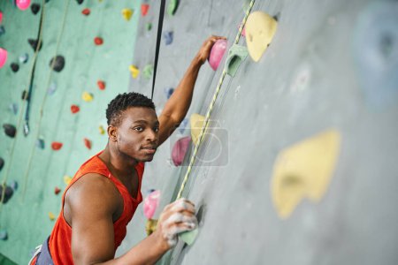 good looking african american man gripping on boulders using safety rope and looking at camera